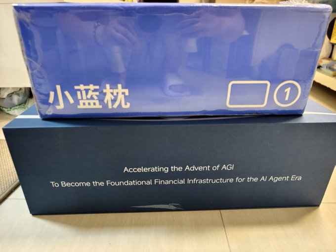 A blue rectangular box labeled "小蓝枕 ①" sits on top of a dark box displaying the English text "Accelerating the Advent of AGI" and "To Become the Foundational Financial Infrastructure for the AI Agent Era," representing the Mid-Autumn Festival gift mentioned in the post from @BAI_AGI.