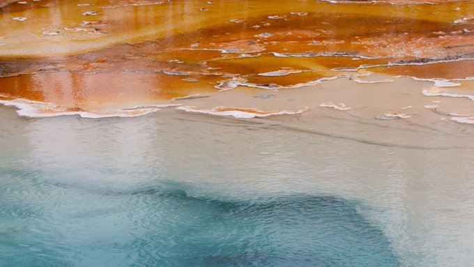 An aerial view of the colorful, layered mineral deposits of a hot spring, showing bands of orange, brown, white, and blue water.