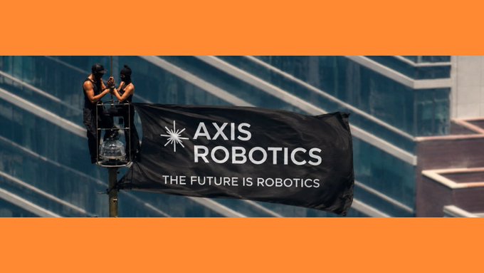 Two people in dark clothing stand on a cherry picker attaching a black banner reading 'AXIS ROBOTICS - THE FUTURE IS ROBOTICS' to a pole, set against a glass building.