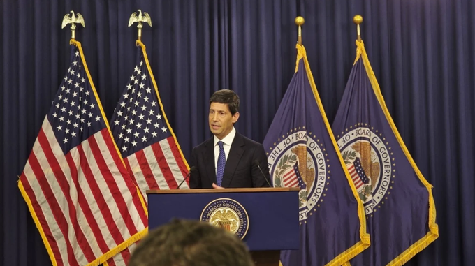 A man in a dark suit speaks at a podium with the Federal Reserve seal, flanked by American flags and Federal Reserve flags against a dark blue curtain backdrop.