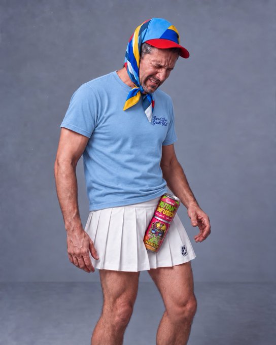 A man in a light blue t-shirt, white pleated skirt, colorful headscarf, and red cap looking down in distress while holding a can labeled 'Nutty Future'.