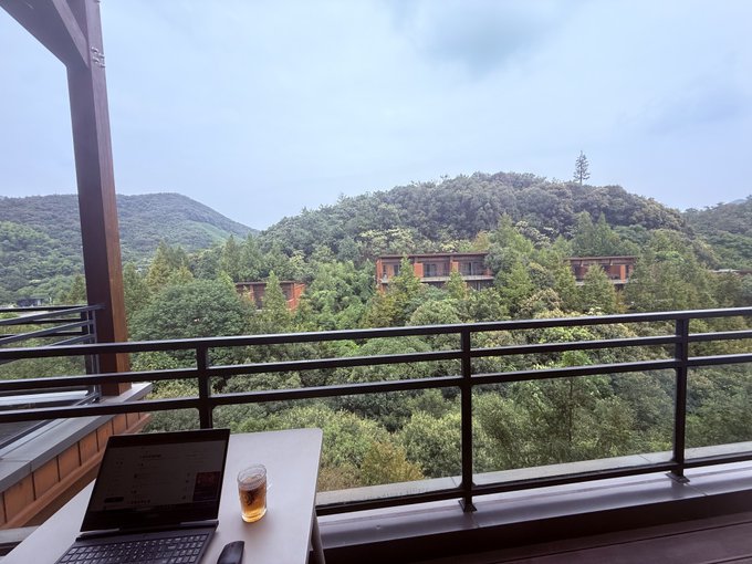 A scenic view from a balcony overlooking lush green mountains and trees, with a laptop and a glass of tea on the table in the foreground.