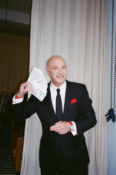 An older bald man in a black suit with a red pocket square smiles while holding up a fan of US dollar bills.