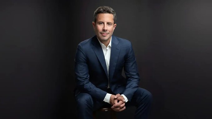 A professional portrait of a smiling man in a dark blue suit sitting on a stool against a dark gray background.