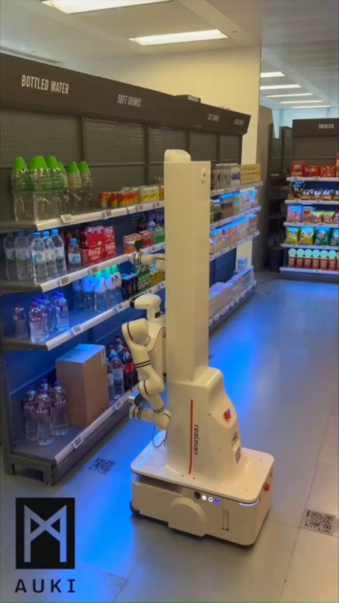 The image features a robot walking down an aisle of a store with shelves filled with various items such as water bottles. The robot appears to be moving at a slow pace, possibly exploring or navigating through the store's layout. The scene suggests that the robot is part of a project named "AUKI" and has been built for $AUKI by a team who have been shipping, building, and executing it.