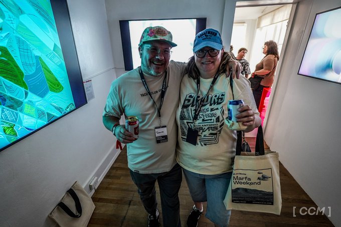 Two conference attendees wearing caps and event shirts pose with drinks and a tote bag; the bag reads “Marta Weekend,” and “[CCM]” appears at the lower right.