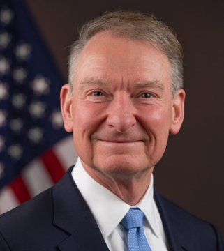 A man with gray hair, wearing a dark suit and blue tie, smiles in front of an American flag, appearing to be an official SEC portrait accompanying news about the SEC's framework for 24/7 tokenized stock trading.