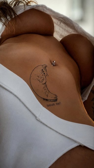 A close-up photograph of a person's torso showing a tattoo of a crescent moon with a face holding a smaller moon, accompanied by the text 'moon me!' and a belly button piercing.