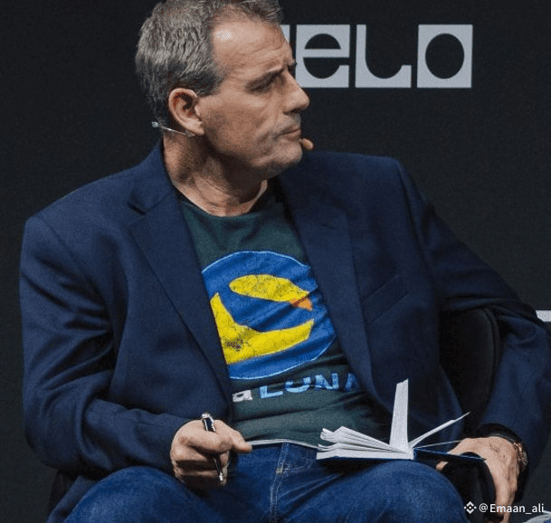 A man in a dark blazer and jeans sits on stage, holding a pen and notepad, wearing a green t-shirt with the original yellow Terra (LUNA) logo. The background shows partial text "elo," likely from a public speaking event, connecting to the post's request for the full image of the classic Terra LUNA OG shirt he is wearing.