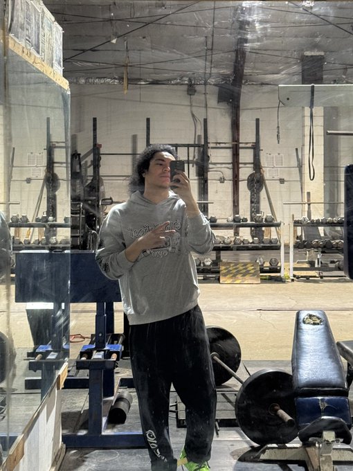 A young man with long dark hair takes a mirror selfie in a gym, wearing a gray sweatshirt and black sweatpants, making a peace sign.