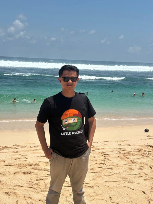 A man stands on a sunny beach wearing sunglasses and a black t-shirt featuring a cartoon character with a face mask and the text "LITTLE MUSTACH," fitting the cheerful "gm" (good morning) greeting. The turquoise ocean and clear blue sky behind him add to the relaxed, morning vibe of the post.