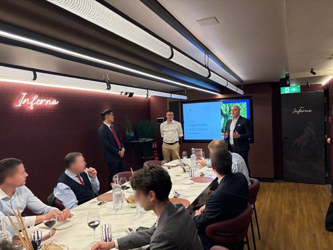 A business meeting or presentation taking place in a venue named 'Inferno', with men seated around a dining table while one presenter stands by a screen displaying 'Digital assets with BITSTAMP'.
