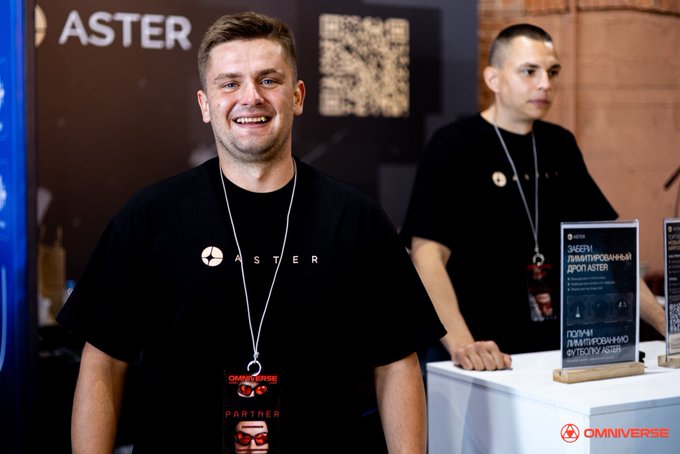 Two men wearing black 'ASTER' t-shirts stand at a conference booth, one smiling at the camera; signage includes 'ASTERO' branding and Cyrillic text mentioning a 'криптовалютный брокер Aster' (crypto broker Aster).