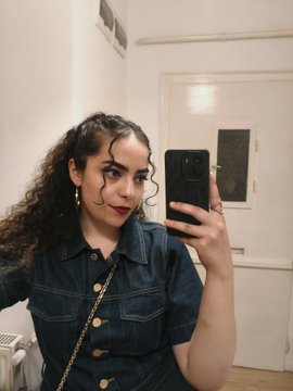 A young woman with curly hair takes a mirror selfie wearing a denim shirt and gold chain strap, holding up her phone. The caption connects to the image by showing the artist pausing for a casual "GM" (good morning) selfie while considering pricing strategies for her 1/1 artworks—debating whether to relist them higher or remint as editions for the same total value.