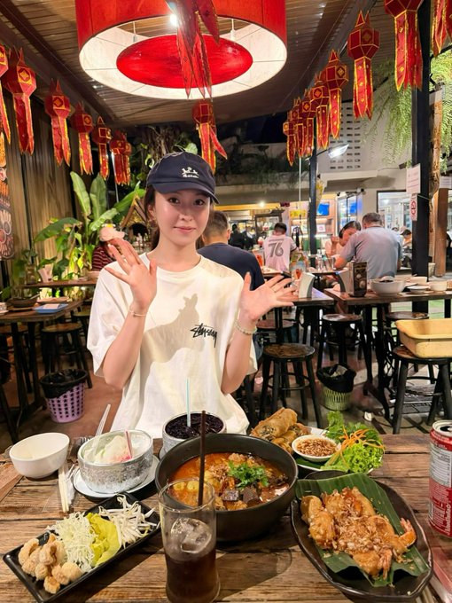 A young woman wearing a black cap and white Stüssy t-shirt poses with her hands up behind a table full of Thai food, including fried chicken, spring rolls, and a hearty bowl of curry, in a restaurant decorated with red Chinese lanterns.