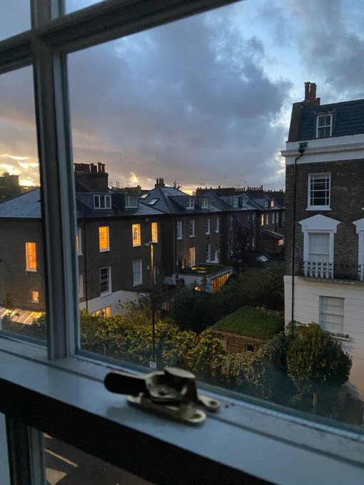A view through a window of a row of brick townhouses at dusk with a cloudy sky, with a window latch visible in the foreground.