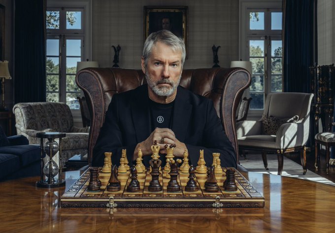 A serious-looking older man with a gray beard, wearing a dark suit, sits with clasped hands behind an ornate wooden chessboard in an elegant, classically furnished room. An hourglass sits beside the board, evoking strategic patience and calculated moves—mirroring the post's question about how Strategy's recent $450 million USD reserve increase will be deployed, likely into Bitcoin.