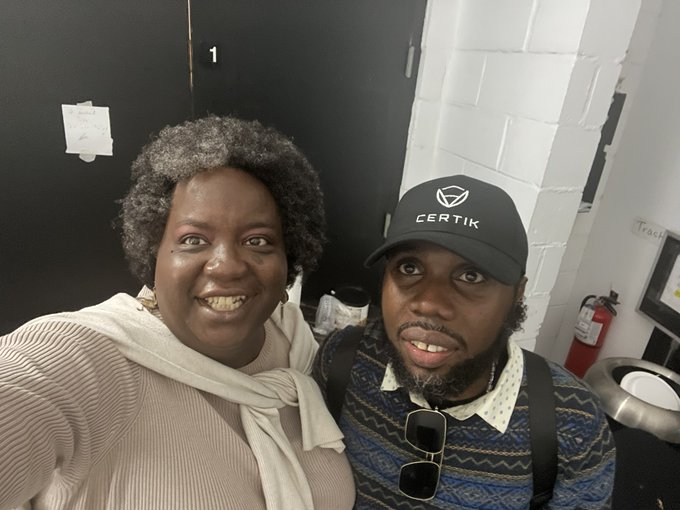 A woman and a man wearing a "CERTIK" branded cap take a smiling selfie together at an indoor event, with a fire extinguisher visible in the background. The image captures an IRL meetup moment referenced in the post, showing the woman posing alongside "the man himself" she has followed online for years.