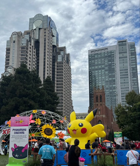 A crowd gathers at what appears to be a Pokémon-themed event in an urban park, featuring a giant inflatable Pikachu and signage reading "Pikachu XP" and "PYUKUMYUKU CUCKING" against a backdrop of towering skyscrapers and a brick church. The image captures the energetic pursuit of a beloved passion—turning fandom dreams into real-world action—echoing the post's message that meaningful goals require more than wishful thinking.