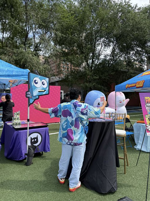 A person in a blue and purple camo shirt holds up a cartoonish blue mascot sign at an outdoor event with booths, pink mascot figures, and tents on a sports field.
