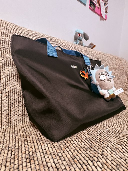 A black HTX-branded tote bag rests on a textured beige sofa, adorned with a Rick and Morty plush keychain and a small fox charm.