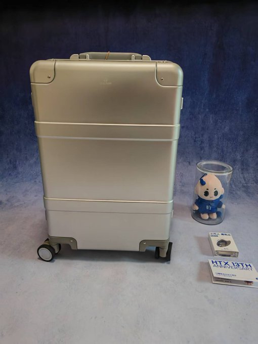 A champagne-colored suitcase stands next to a small blue and white mascot figurine in a clear cup and two packs of Panasonic 'Ni-MH' AAA batteries on a blue fabric background.