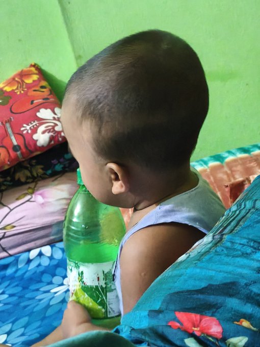 A young child with a shaved head drinking from a green plastic Sprite bottle while sitting on a bed with blue floral bedding.