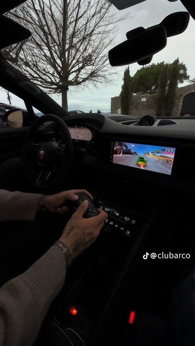 A first-person view from inside a luxury car (Porsche steering wheel) where a person with tattooed hands is playing a video game on the dashboard screen; text reads '@clubarco'.