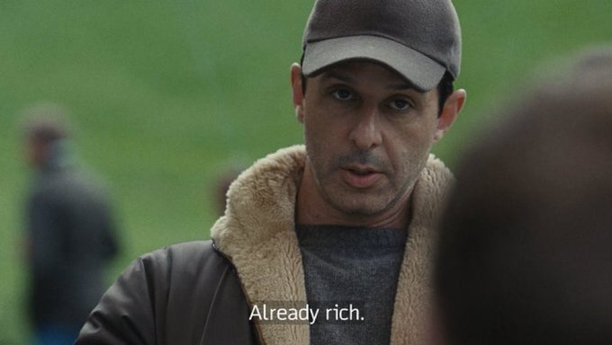 John Travolta as Vincent Vega in *Pulp Fiction* wears a cap and shearling-lined jacket while delivering the line "Already rich," humorously illustrating a trader's motive of profiting from pump-and-dump schemes regardless of price.