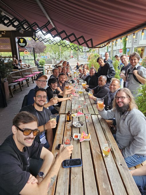 A large group of people sits at a long wooden picnic table on an outdoor patio, raising glasses of beer in a toast.