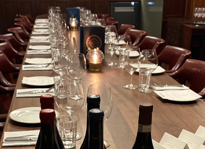 An elegantly set long wooden dining table prepared for a formal dinner, featuring wine bottles, multiple wine glasses, menus, and lit candles.