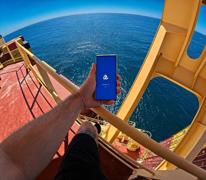 First-person view from a high platform over the ocean, holding a smartphone displaying a blue screen with a sailboat logo and the text 'tether.io'.
