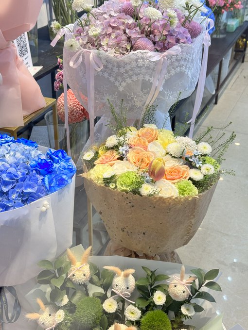 The image shows a display of floral arrangements, including bouquets featuring blue hydrangeas, orange and yellow roses wrapped in brown paper, and decorative arrangements with bunny-shaped decorations, all set in what appears to be a flower shop.