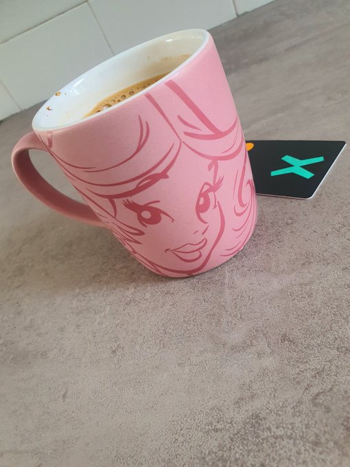 A pink ceramic mug featuring an anime-style character design, containing coffee, sitting on a concrete surface next to a black card with a teal 'X' logo.