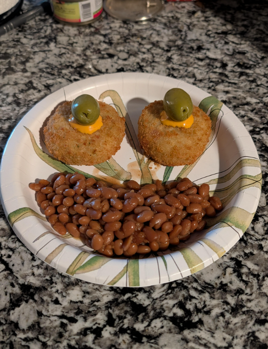 The image features two food items, a plate with beans and a small piece of food shaped like an eye, placed on a table. The beans are browned and appear to be seasoned, while the eye-shaped food item has been made into a face using green olives as eyes. The background is blurred, but it seems to be a kitchen or dining area where the plate with the food items is located.