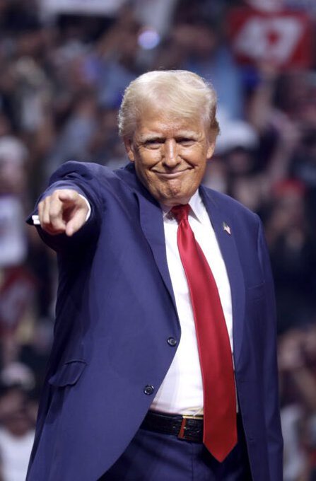 Donald Trump is shown in a blue suit with a red tie, smiling and pointing forward at what appears to be a rally, with a crowd and a Canadian flag visible in the background. The image relates to the post about the "$TRUMP" meme cryptocurrency token, illustrating the political figure after whom the token is named and whose namesake is being used to promote its dramatic launch returns.