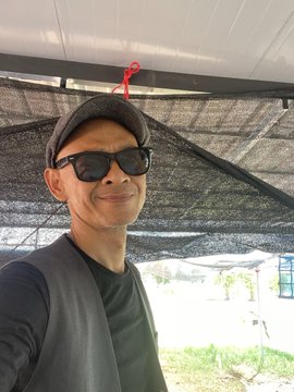 The image features a man wearing sunglasses, a black shirt, and a gray vest standing under a tent or canopy. The man has his hand raised for the camera, capturing a moment of him posing with the tent behind him. The caption of the image reads "Just me & my Rider Wherever I go he go My Rider @SpaceRidersXYZ". This suggests that the man is likely sharing this photo on social media or with friends to showcase an interesting location or event in his life.