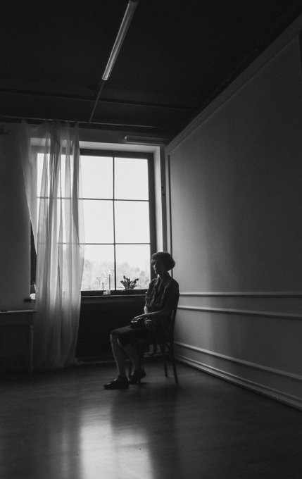 A black and white photograph of a person sitting on a chair by a bright window with sheer curtains in a sparsely furnished room.