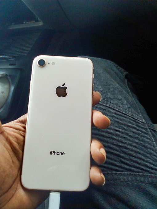 A hand holds a white Apple iPhone showing the back with the Apple logo and 'iPhone' text, with a charging cable connected.
