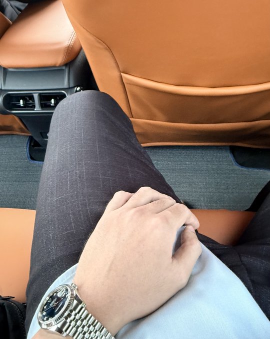 A man wearing a dark checkered suit and a silver Rolex watch sits in the back seat of a car with tan leather interior, his hand resting on his knee.