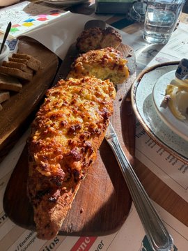 A close-up photograph of cheesy garlic bread served on a wooden board with a knife, accompanied by a plate of food and a water glass at a restaurant table.