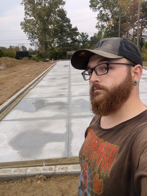 A bearded man with glasses and a cap takes a selfie while standing next to a freshly poured concrete foundation slab at a construction site.
