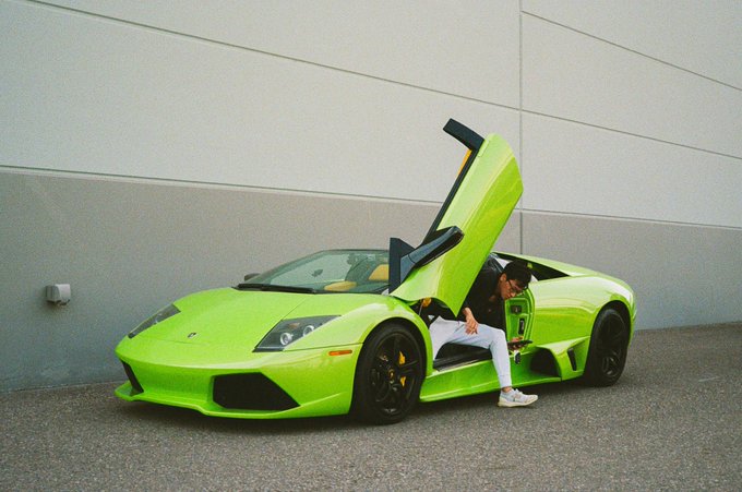 A bright green Lamborghini Murcielago with its scissor door open, parked against a beige wall with a person seated inside.