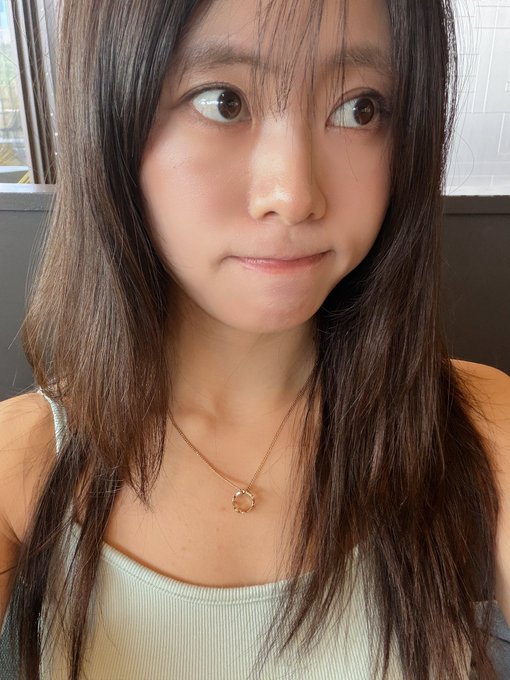 A close-up selfie of a young woman with long dark hair wearing a light green tank top and a delicate gold necklace with a clover pendant.