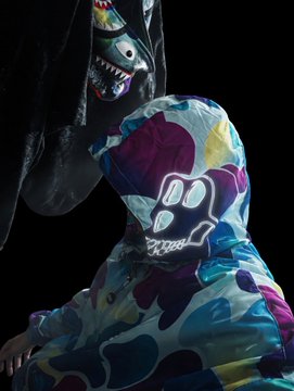 The image features a person wearing a hoodie with a camouflage pattern, which has a skull design on the front. The hoodie appears to be made of a soft material like fleece or velvet. In the background, there are two other people present - one standing and another sitting down. They seem to be engaged in conversation or observing something happening nearby. The post that accompanies this image is titled "BAPE × BAYC Beach Club Edition Wearing @BoredApeYC feels so good. Grail #3 Secured". This post seems to be promoting a beach club event, possibly an outdoor gathering or party where people can enjoy the company of others and the beautiful surroundings while wearing their hoodies with unique designs.