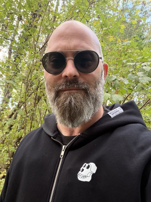 A bald man with a gray beard wearing round sunglasses and a black hoodie with a small dog skull logo poses for a selfie in front of green foliage.