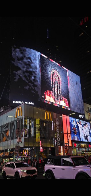 A nighttime cityscape featuring brightly lit billboards and signs, including a McDonald's logo, a 'SARA KIAN' advertisement, and a 'GIFTS' shop, with cars visible on the street below.