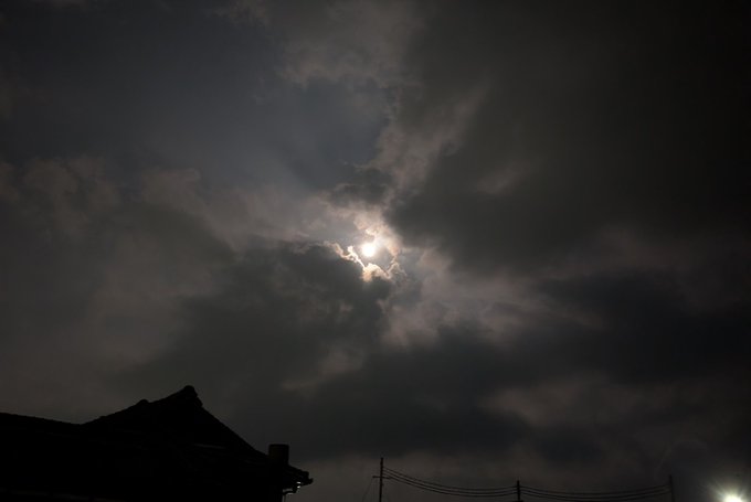 The moon glows brightly through a veil of scattered clouds in a dark night sky, with the silhouetted roof of a building visible in the lower portion of the frame, capturing the serene beauty referenced in the post's caption "The moon is beautiful, isn't it" (#STEPN).
