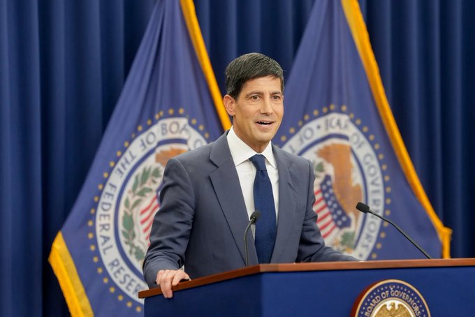 A man in a navy suit and tie speaking at a podium, standing in front of a blue flag with the seal of the Federal Reserve Board of Governors.