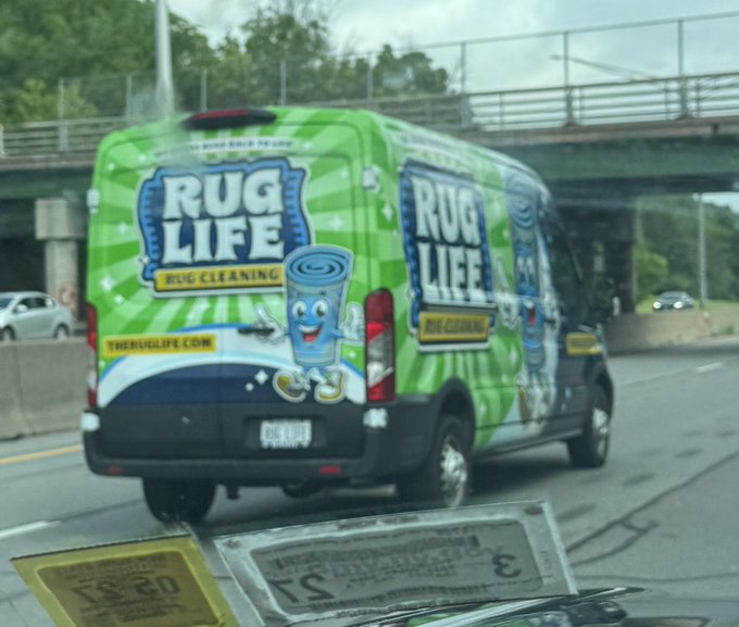 A "Rug Life" rug cleaning van is shown driving on a highway, with the playful slogan clearly visible. The image relates to the post as a humorous play on "rug pull," a crypto scam where developers abandon projects after taking investors' money, visually personifying the concept as a rug literally being taken away.